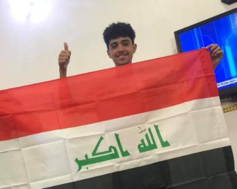 A boy poses for a photo, giving a thumbs up while holding a red, white and black flag.