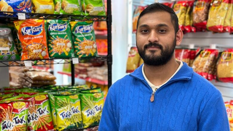 A man stands looking at the camera wearing a blue zip up sweater with snacks from India and Pakistan in packets on the wall behind him.