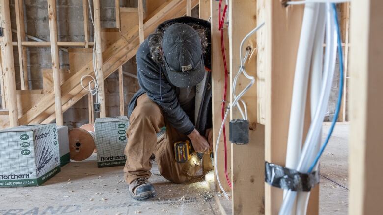 A construction worker is crouched down inside of a structure that looks incomplete. They are holding a drill and are in the process of using it.