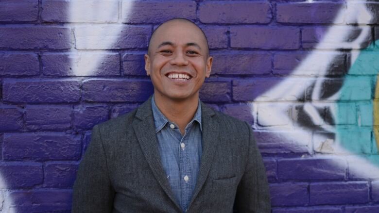 A smiling bald man in front of a purple brick wall and wearing a grey blazer and jeans shirt