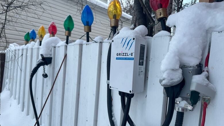 A white electrical box and some thick black cords attached to a snow-covered white fence. The fence has Christmas lights on top of it.