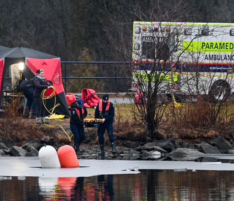 Police divers walk towards a river in winter with a small robot they'll use to search it.