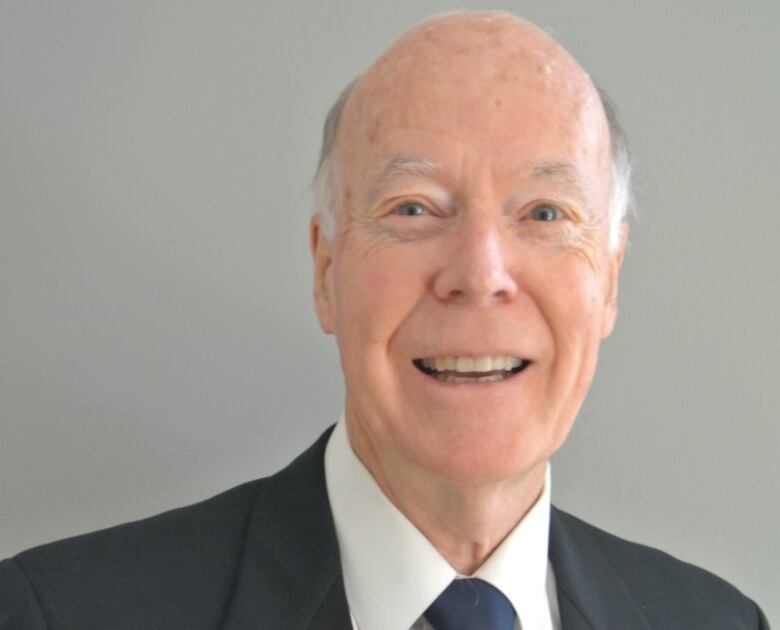 A man in a black suit wearing a blue tie smiles at the camera.