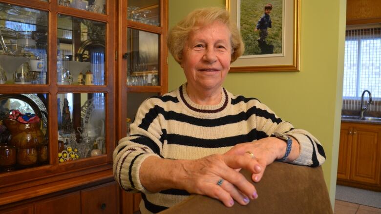 A senior woman in her friend's dining room resting her hands against the back of a chair. 