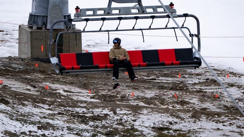 A skier on a ski lift with brown soil and dirt below him.