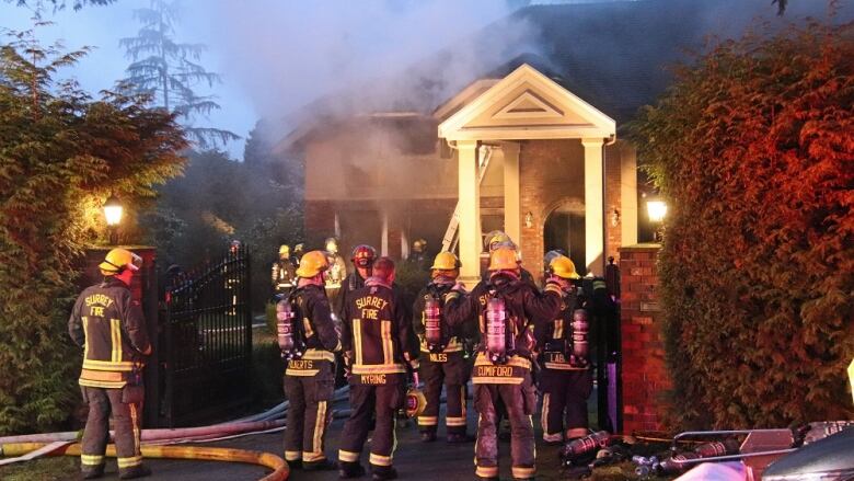 More than a dozen firefighters stand outside a home with smoke billowing out of it.