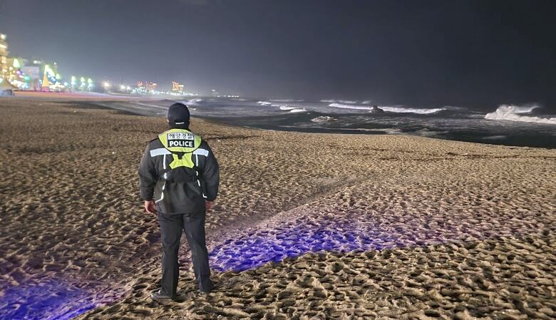 A man wearing a police uniform stands on a beach at nighttime.