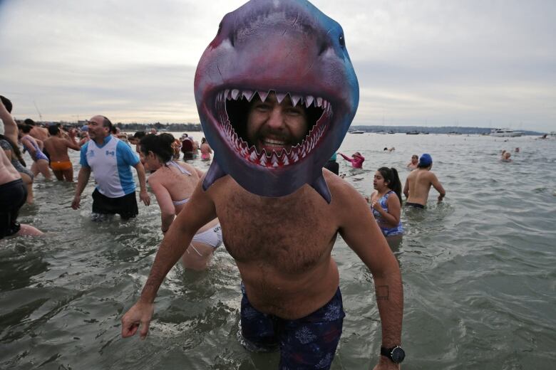 A man wearing a shark's head costume smiles as he emerges from beachwater, with lines of other people behind him taking a dip.