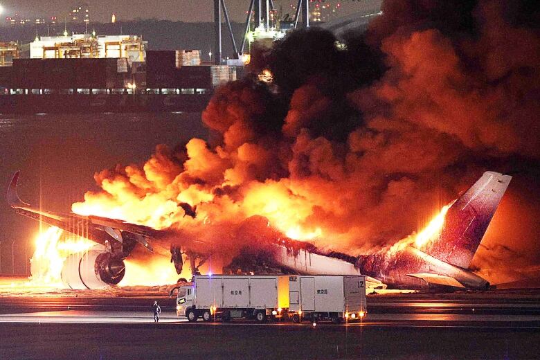 A person and a truck are dwarfed by a large passenger plane completely engulfed in flames.