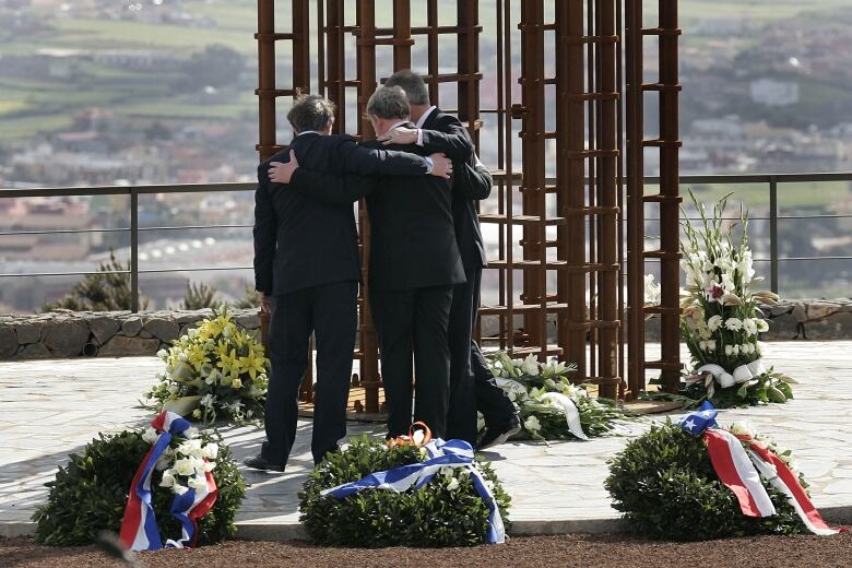 Three people stand arm in arm near commemorative flowers at the base of a large monument.