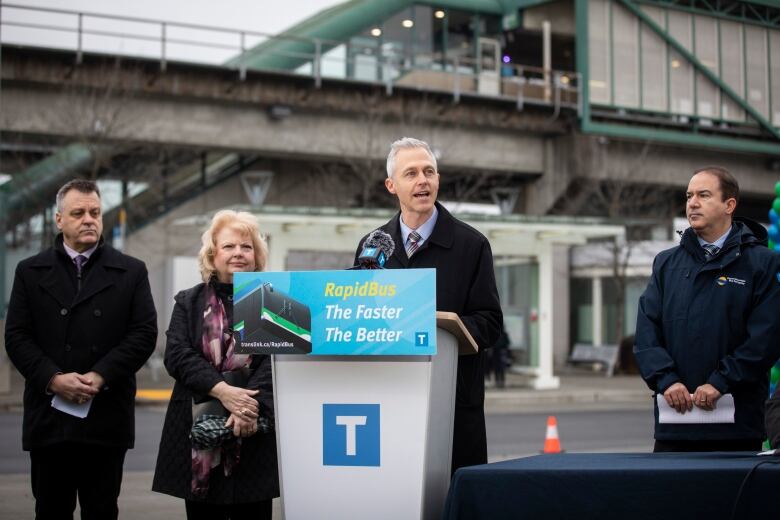 A man in a blue shirt, tie and black blazer jacket is seen speaking at a podium, on which a sign reading 'RapidBus, The Faster, The Better' is mounted, along with a logo bearing the letter 'T'. Standing behind him, left to right, is a man in a black blazer, a woman in a black jacket and purple scarf, and a man in a hooded jacket. 