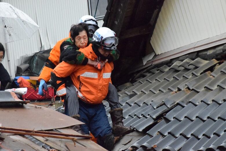 A woman clasps her arms around a man in an orange vest and helmet as she rides on his back amid the ruins of a building.