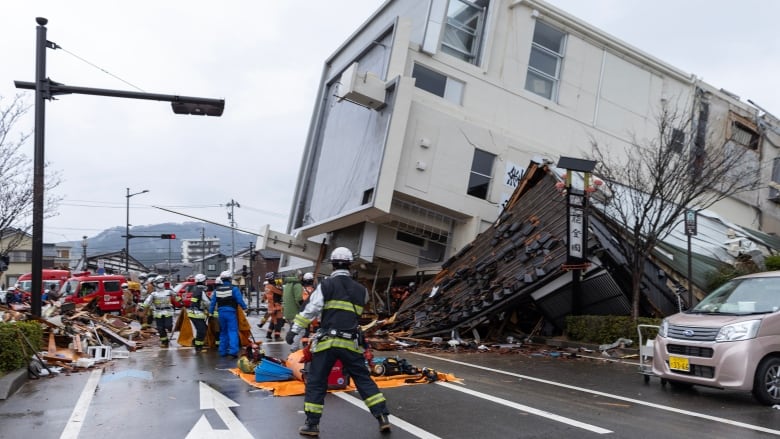 A largely intact multi-storey building lies on its side. People in helmets stand nearby in the street.