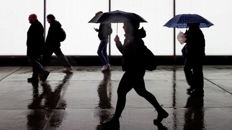 Five people walk down a rainy, wet sidewalk against a brightly lit building at night. Two are holding umbrellas.