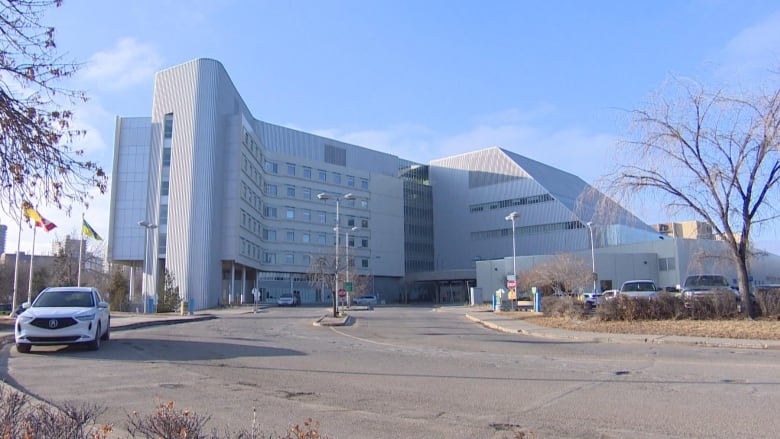 An outdoor shot of a hospital, with cars parked at the edge of the sidewalk and flags waving outside.