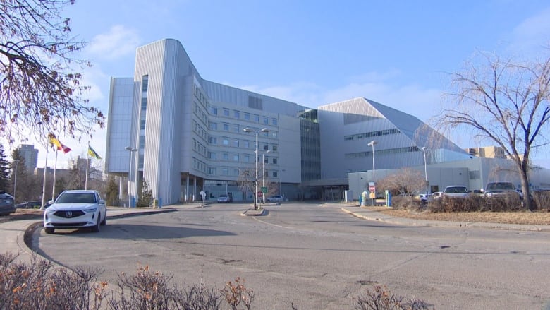 An outdoor shot of a hospital, with cars parked at the edge of the sidewalk and flags waving outside.