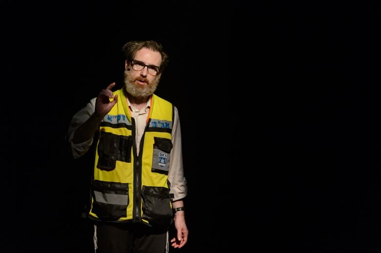 A man wearing a yellow jacket with blue writing in Hebrew is standing in a spotlight speaking. 