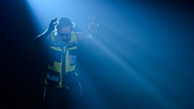 A man wearing a yellow jacket is bent over with his hands stretched out in front. He's covered by a blue spotlight. 