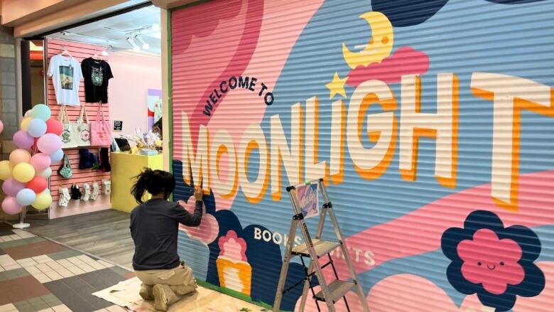 a woman paints a colourful shop sign that says 