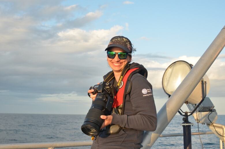 Young woman wearing sunglasses and ball cap stands on boat in ocean with large camera around her neck.