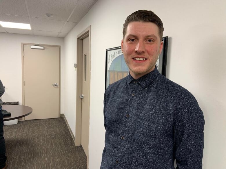 A man in a buttom up blue dress shirt stands in front of a white wall. 