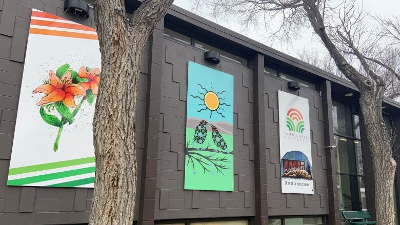 Bright, colourful Indigenous paintings line a brown wall. Trees line the wall as well. 