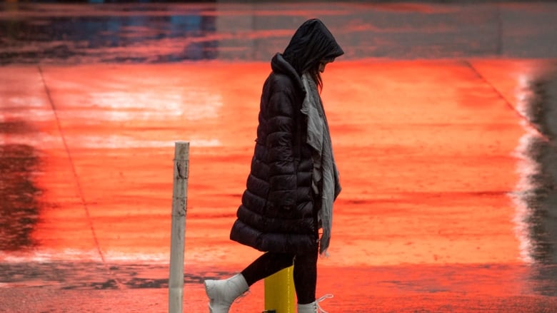 A woman in a long jacket walks down a wet street with her head down.  