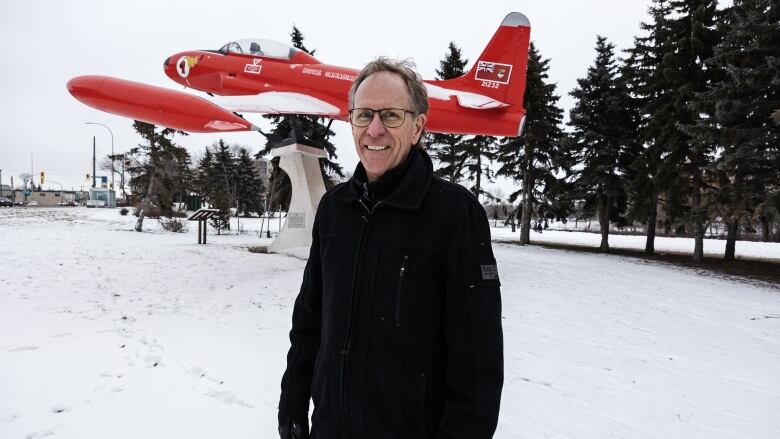 A man stands in a snowy field in front of a monument featuring a small red airplane.