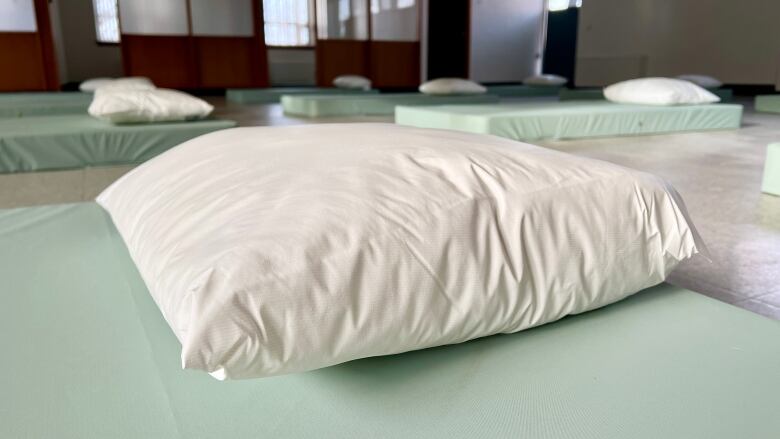 A closeup shows a pillow on a mattress in a room, with several other mattresses neatly arranged on the floor in the background.