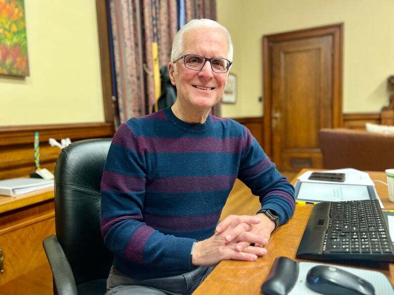 A smiling man in a sweater sits at a desk in an office.