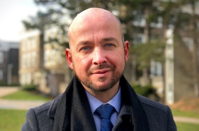 A man in a dark suit with a blue tie looks at the camera