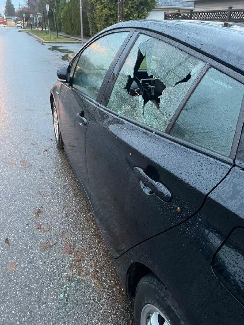 A car with its rear window smashed sits on a rainy street.