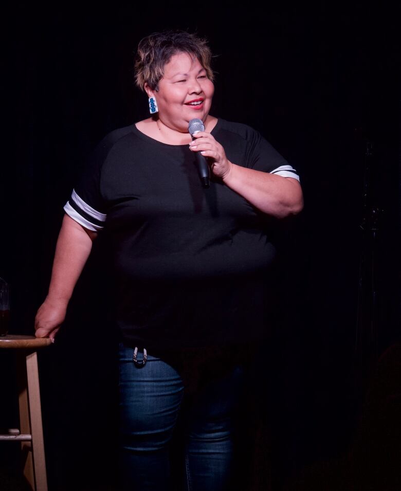 Annie Brass with a microphone and near a stool in a stand up comedy setting