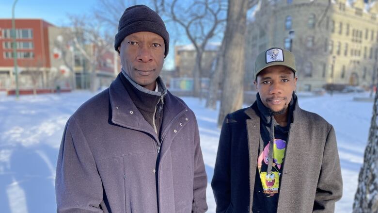 Two men wearing coats and hats stand outside and look into the camera.