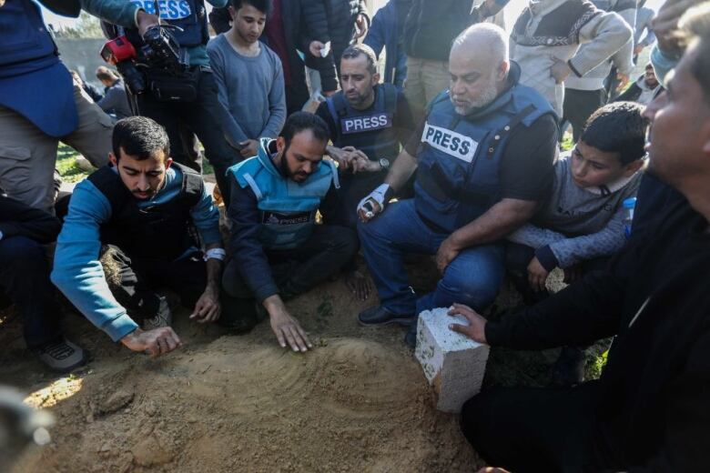 Men in press vests surround the dirt and cement block headstone marking a newly dug grave