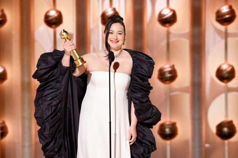 A woman wearing a white gown and a black shawl holds a golden statuette in one hand while speaking into a microphone.