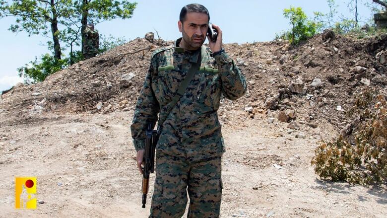 A man in combat fatigues and carrying a weapon while speaking into a device.