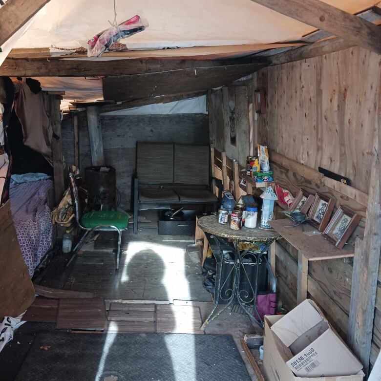 The inside of a shack, with a bed, table and chairs and the shadow of a woman standing in the doorway.