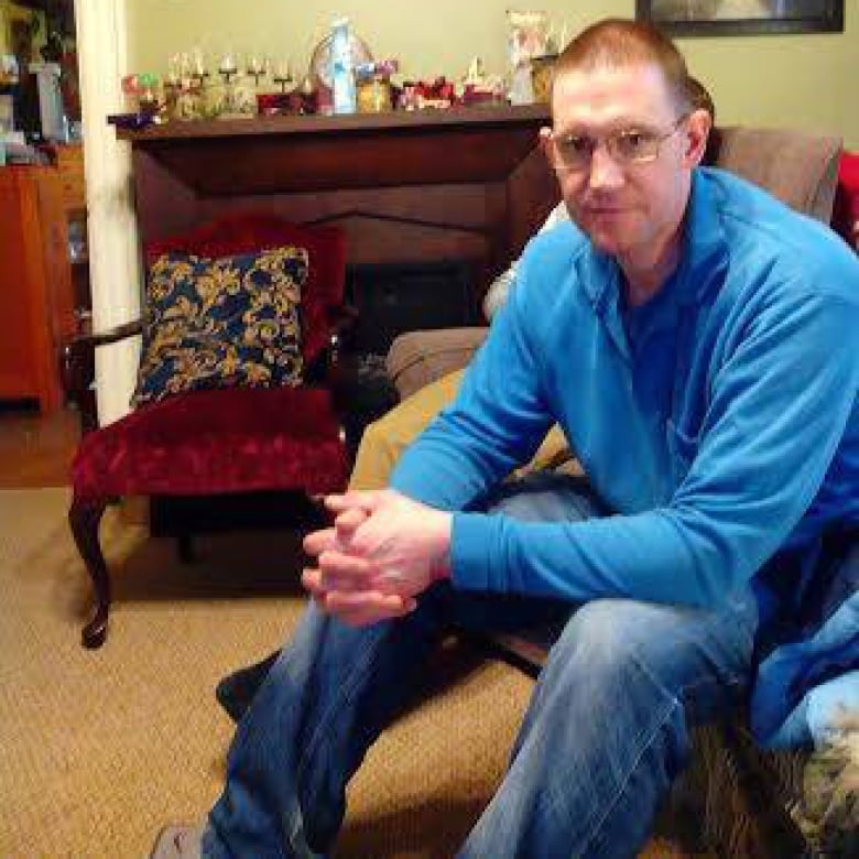 A man with close-cropped hair and glasses, wearing a blue shirt and jeans, sitting in a chair in a living room.