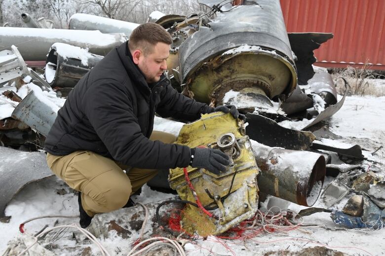 A bearded man wearing gloves crouches on a snowy filed to hold and examine a large piece of metal that contains wiring.