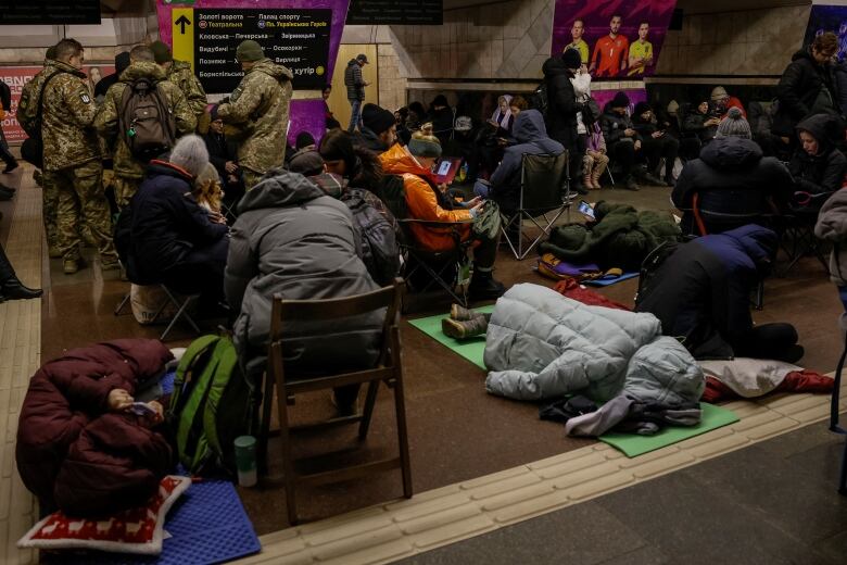 Dozens of people in heavy coats, as well as military personnel in camouflage gear, huddle at an indoor location.