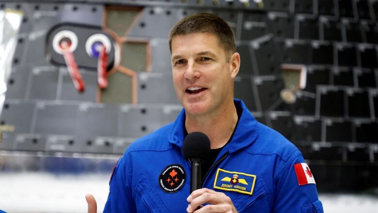 A male astronaut in a blue jumpsuit holds a microphone and speaks in front of a grey spacecraft.