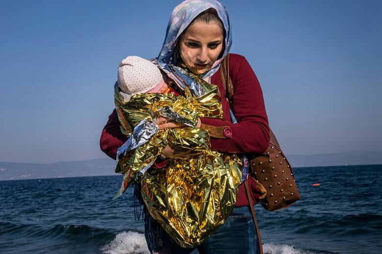 A woman wearing holds her baby in a reflective blanket by the sea.