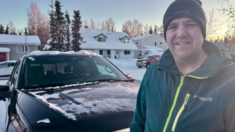 A man in a green coat and a black hat standing in front of a dark coloured truck.