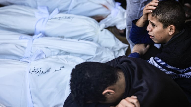 A boy buries his head in his arms next to another boy, who is crying, as they sit next to a row of white body bags.