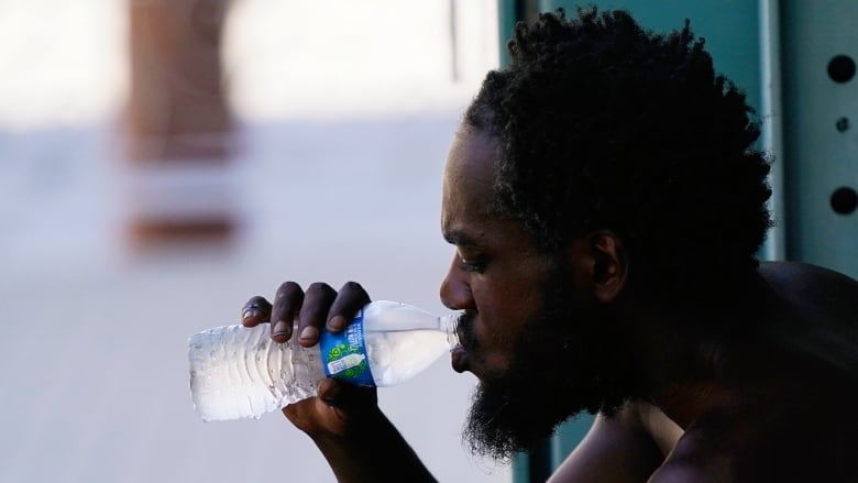 A shirtless man in profile is silhouetted drinking a bottle of water while crouched or leaning against a building.