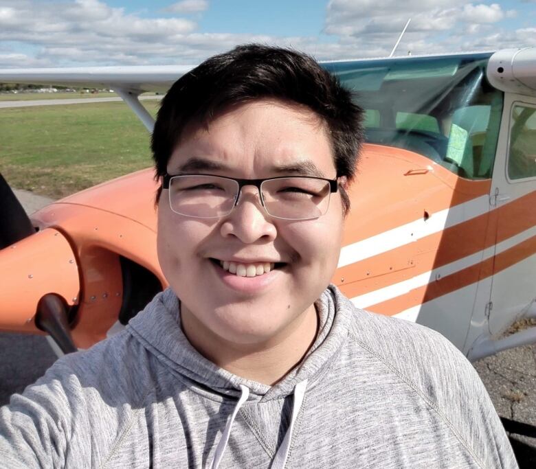 A young man wearing black-framed glasses poses for a selfie in front of a small orange and white airplane