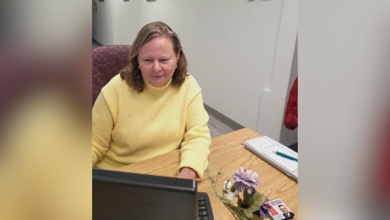 a woman in a yellow shirt works at a computer. There is also a flower and small pamphlets on her desk. 