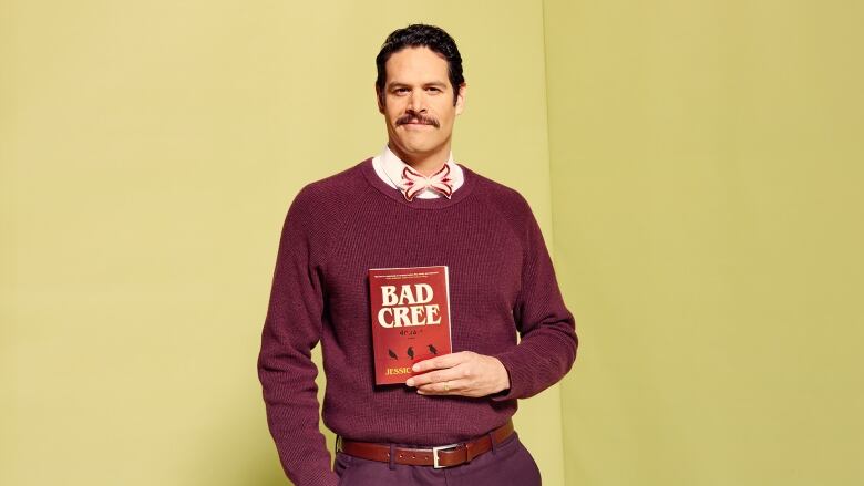 A Cree man with a moustache holds up a red book. He is wearing a maroon outfit. 