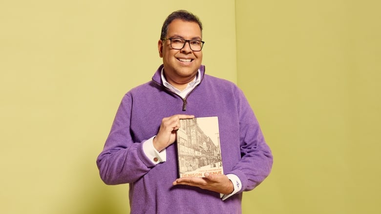 A South Asian man wearing glasses and a purple quarter zip sweater smiles at the camera while holding a book. 